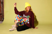 A woman sitting on a yellow-green studio floor wearing a burgundy graphic sweatshirt with a colorful duck design and the word 'Lucky', a bright yellow beanie, a multicolor quilted skirt, black pants, and beige sneakers.