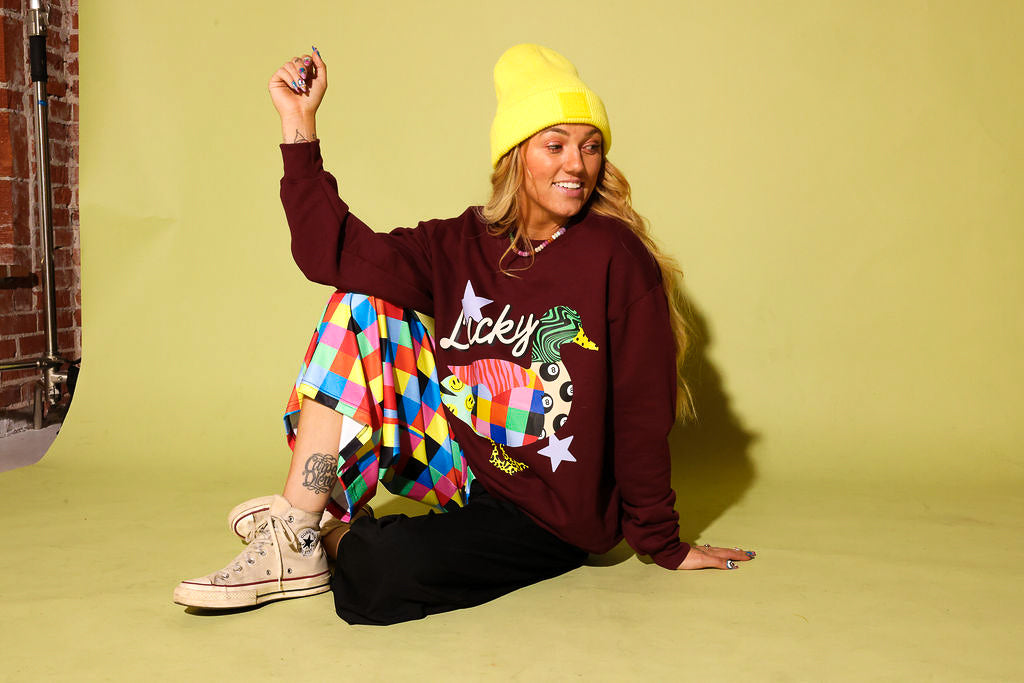 A woman sitting on a yellow-green studio floor wearing a burgundy graphic sweatshirt with a colorful duck design and the word 'Lucky', a bright yellow beanie, a multicolor quilted skirt, black pants, and beige sneakers.