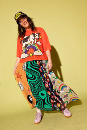 A person modeling a vibrant orange graphic t-shirt with a cow and rainbow print, paired with a highly patterned multicolor maxi skirt featuring psychedelic swirls, cartoon faces, and stars, set against a solid pale yellow-green studio background.