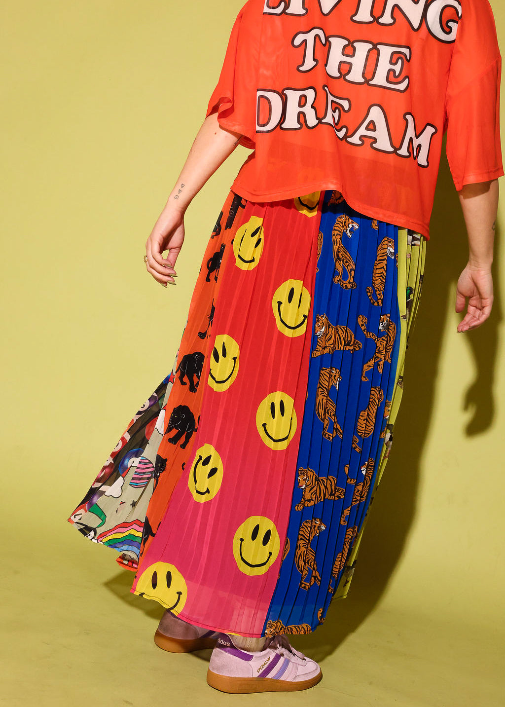 A person wearing a bright orange graphic T-shirt and a long pleated skirt with multiple bold panels, including yellow smiley faces, black-and-white animal prints, and a blue section, standing against a pale yellow-green studio background.