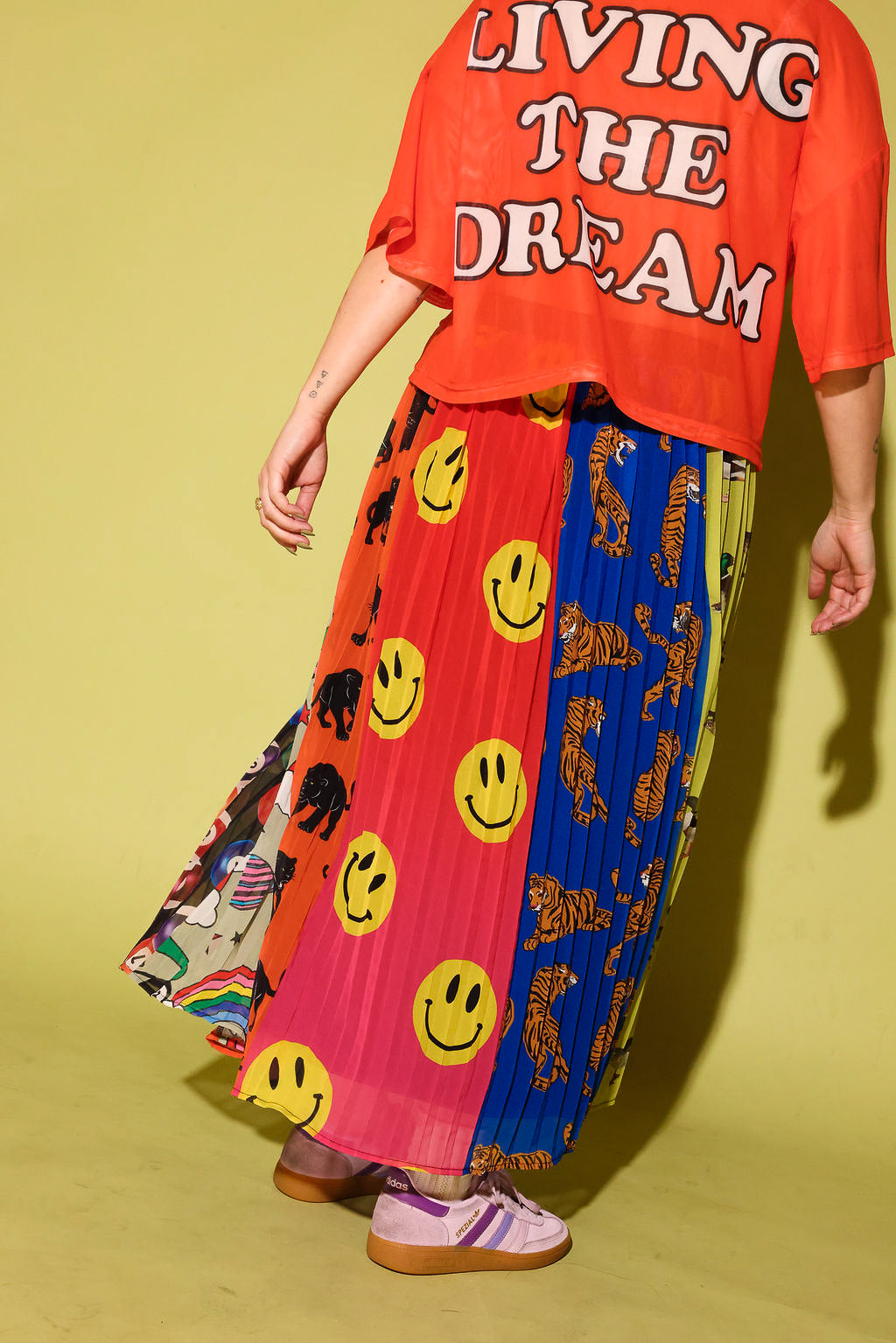 A person wearing a bright orange graphic T-shirt and a long pleated skirt with multiple bold panels, including yellow smiley faces, black-and-white animal prints, and a blue section, standing against a pale yellow-green studio background.