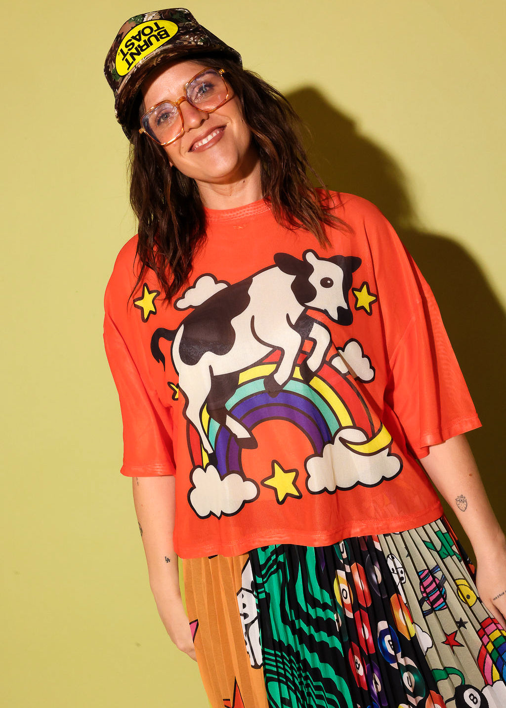 A person wearing an oversized bright orange graphic T-shirt with a cow riding a rainbow design, paired with a patterned skirt and a cap, standing against a solid pale yellow-green background.