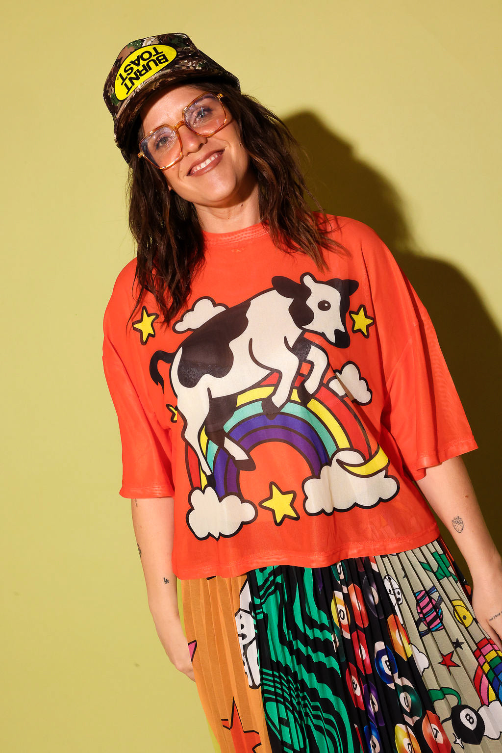 A person wearing an oversized bright orange graphic T-shirt with a cow riding a rainbow design, paired with a patterned skirt and a cap, standing against a solid pale yellow-green background.