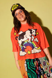 A person wearing an oversized bright orange graphic T-shirt with a cow riding a rainbow design, paired with a patterned skirt and a cap, standing against a solid pale yellow-green background.