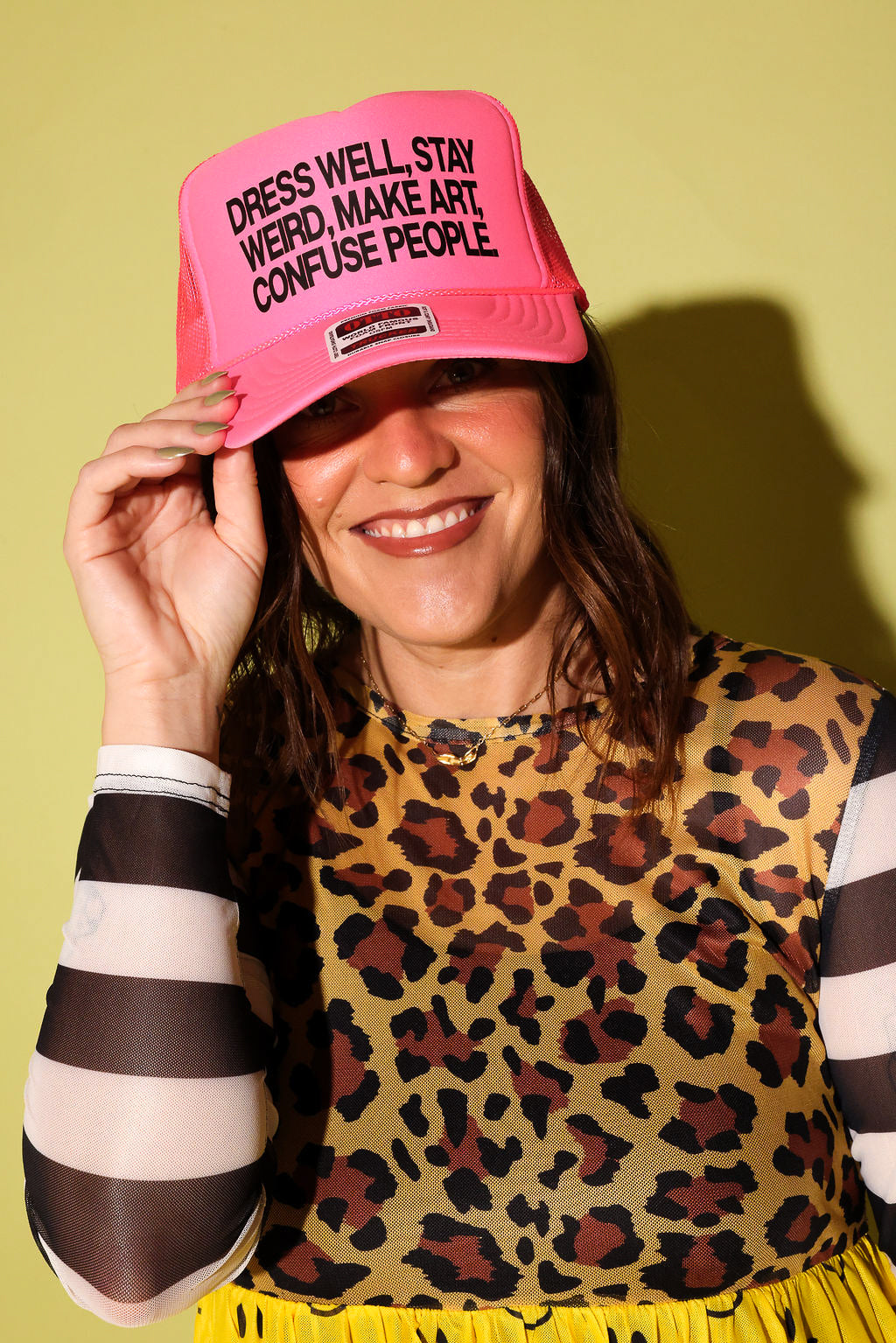 A woman wearing a bright pink trucker hat with black text that reads 'DRESS WELL, STAY WEIRD, MAKE ART, CONFUSE PEOPLE' and a leopard-print top, posing against a solid yellow-green background.