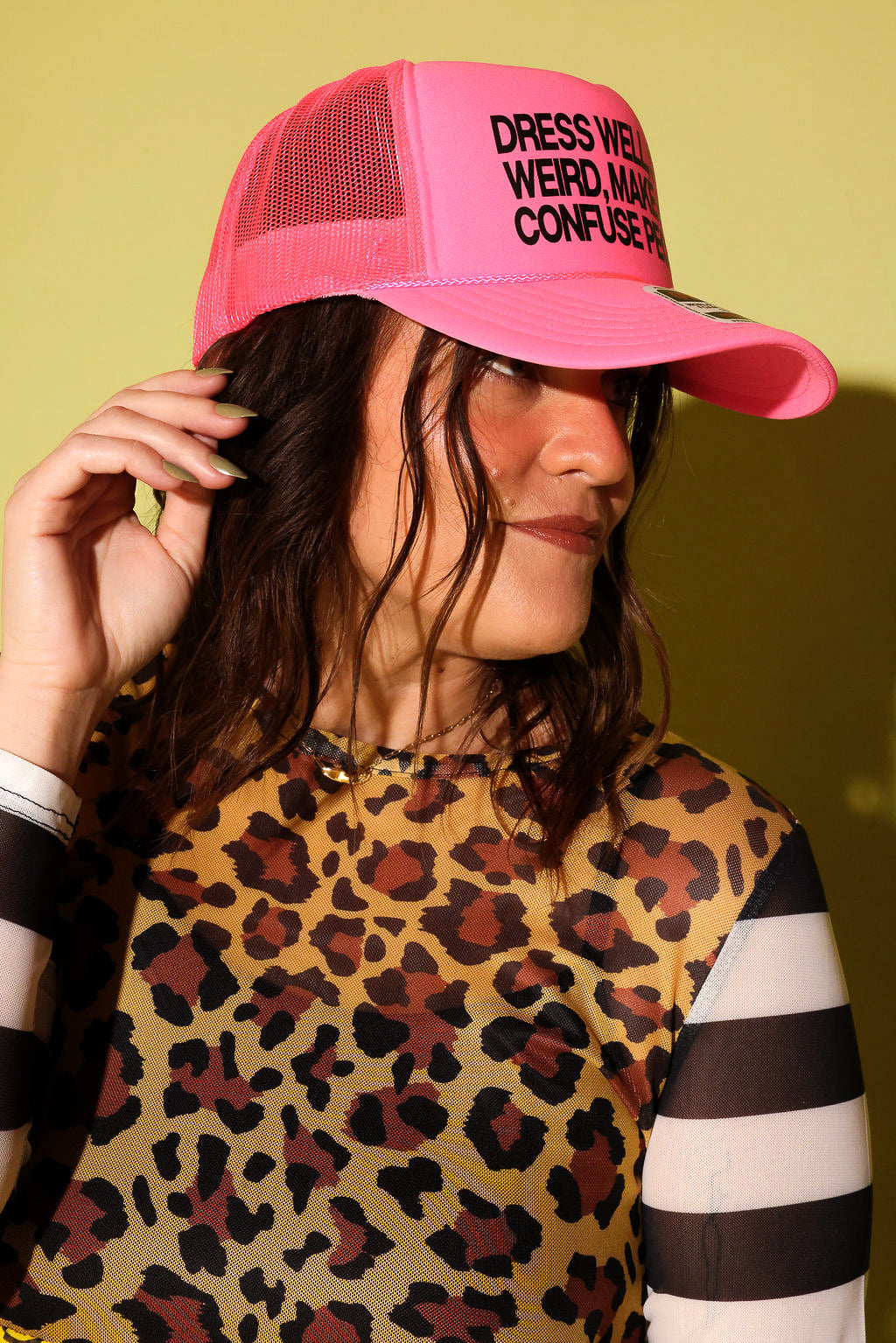 A woman wearing a bright pink trucker hat with black text that reads 'DRESS WEIRD WEIRDLY CONFUSE PEOPLE' and a leopard-print top, posed against a solid yellow-green background.