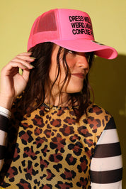 A woman wearing a bright pink trucker hat with black text that reads 'DRESS WEIRD WEIRDLY CONFUSE PEOPLE' and a leopard-print top, posed against a solid yellow-green background.