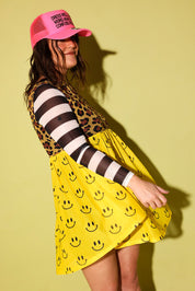 A woman wearing a bright yellow smiley-face printed dress with a leopard-print and black-and-white striped bodice, paired with a pink trucker hat, posed against a solid pale yellow-green background.