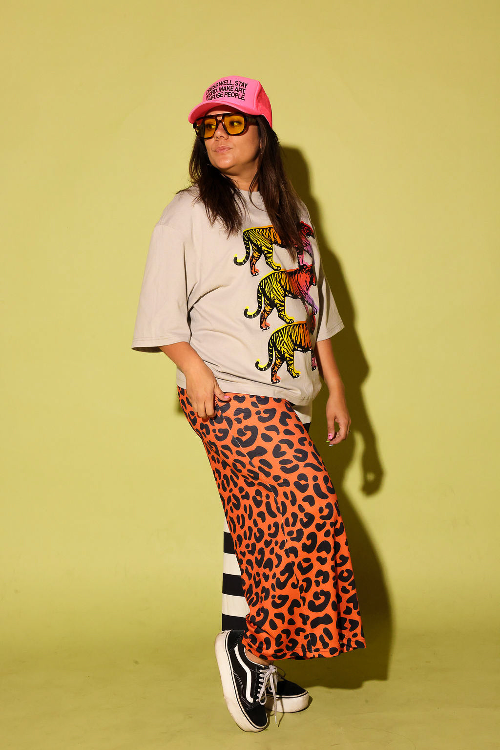 A person wearing a long split-design pants with one side in orange leopard print and the other in black and white stripes, paired with a cream top with tigers and black slip-on shoes, standing sideways in front of a bright yellow wall