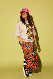 A person wearing a long split-design pants with one side in orange leopard print and the other in black and white stripes, paired with a cream top with tigers and black slip-on shoes, standing sideways in front of a bright yellow wall