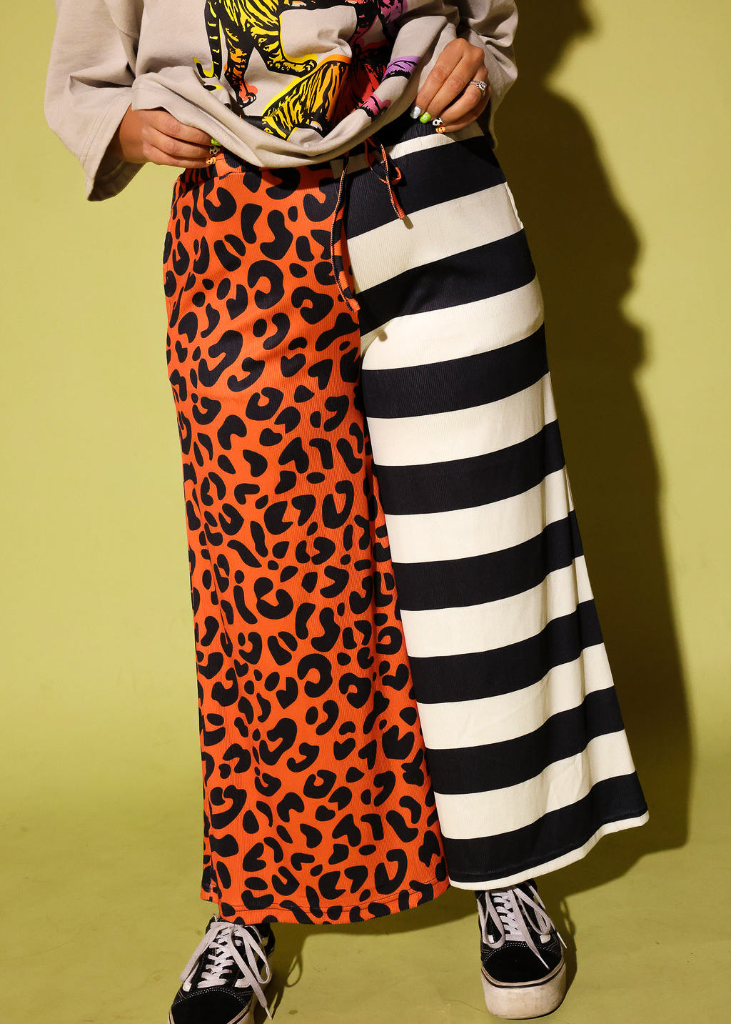 A person wearing a long split-design pants with one side in orange leopard print and the other in black and white stripes, paired with a cream top and black slip-on shoes, standing in front of a bright yellow wall