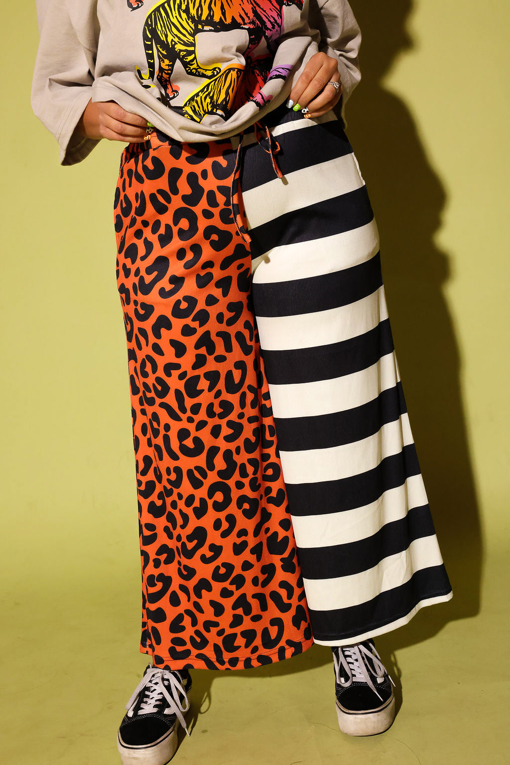 A person wearing a long split-design pants with one side in orange leopard print and the other in black and white stripes, paired with a cream top and black slip-on shoes, standing in front of a bright yellow wall