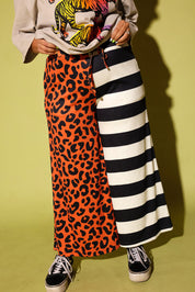 A person wearing a long split-design pants with one side in orange leopard print and the other in black and white stripes, paired with a cream top and black slip-on shoes, standing in front of a bright yellow wall