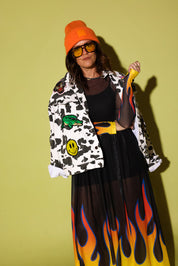 A model wearing a vibrant orange beanie, yellow-tinted sunglasses, a black sheer top, a black skirt with a yellow, orange, and black flame pattern, and a white cow-print jacket with colorful patches, posed against a solid olive-green background.
