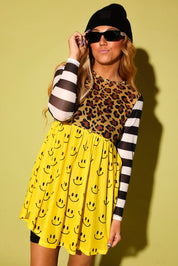A woman modeling a yellow smiley face print mini dress with a leopard print bodice and black-and-white striped sleeves, styled with a black beanie, black sunglasses, and black leggings against a yellow-green background.