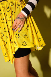 A close-up of a person wearing a bright yellow mini skirt with an all-over black smiley face pattern, styled with a black and white striped long-sleeve top and black fitted shorts against a yellow wall.