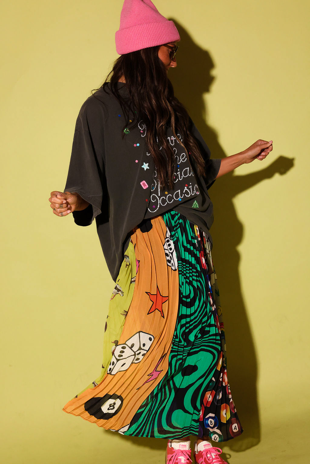 A person wearing a bright pink beanie, a dark oversized graphic T-shirt, and a long colorful skirt with psychedelic patterns, standing against a yellow-green studio background.