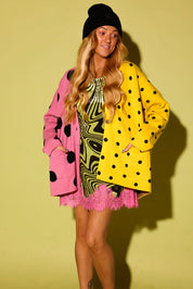 A woman modeling a vibrant, multi-colored outfit consisting of a bright yellow cardigan with black polka dots, a pink cardigan with black polka dots, a pink ruffled mini skirt, and a black beanie, set against a solid pale yellow-green studio background.
