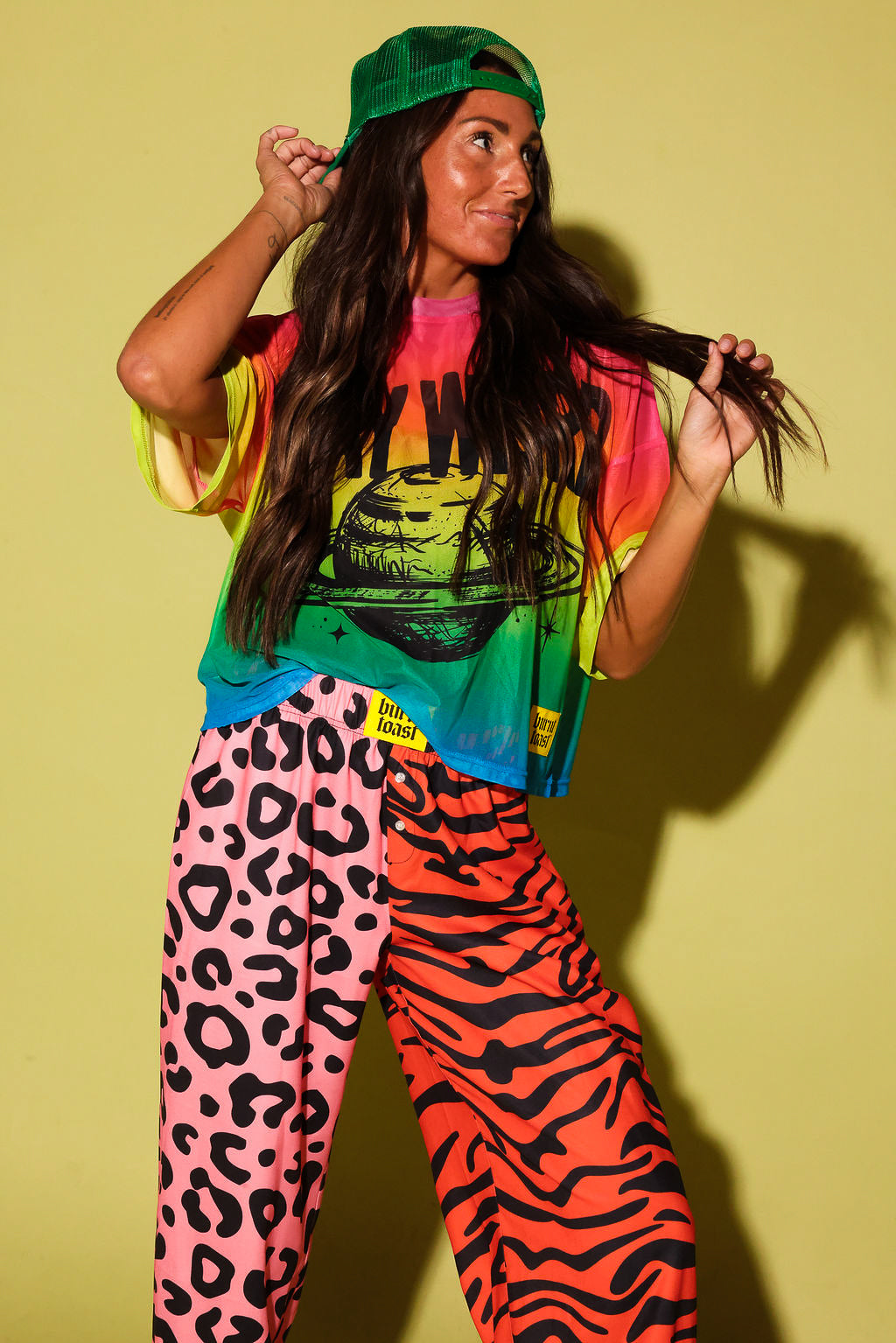 Close up of a model wearing orange patterned jogger pants and yellow burnt toast tag, a colorful oversized mesh tee, and a green trucker hat, against a pale yellow background.