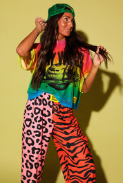 Close up of a model wearing orange patterned jogger pants and yellow burnt toast tag, a colorful oversized mesh tee, and a green trucker hat, against a pale yellow background.