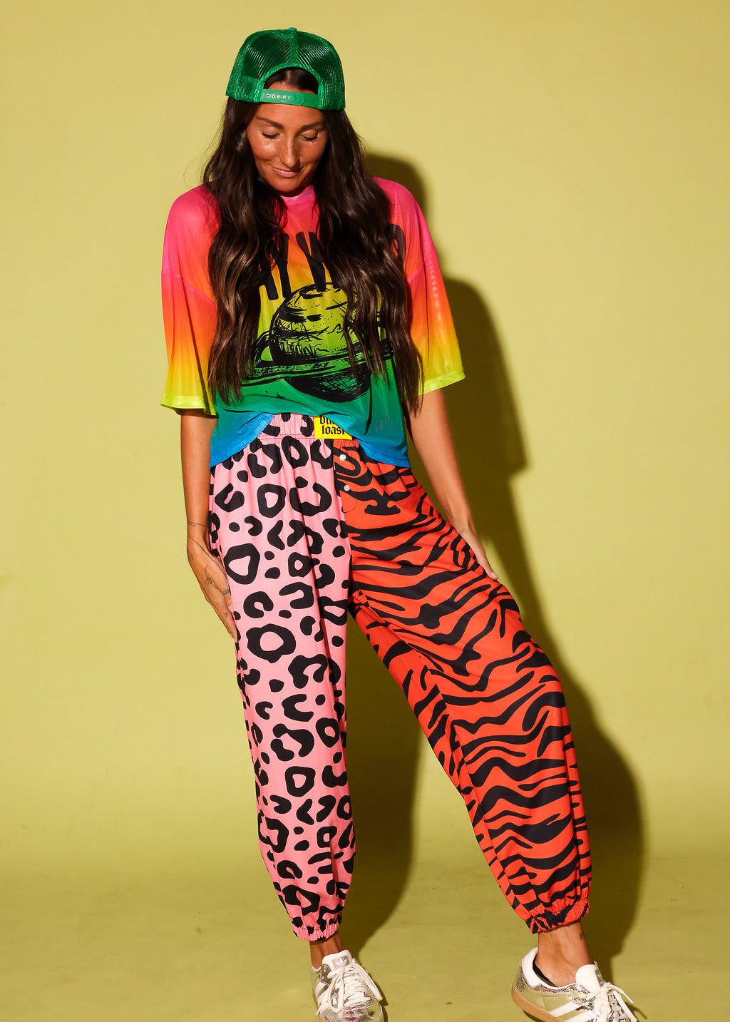Model wearing a colorful oversized mesh tee, pink and orange patterned jogger pants, green trucker hat, and sneakers against a pale yellow background.