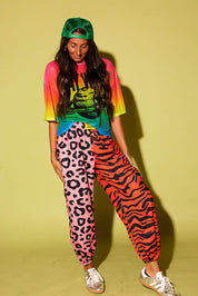 Model wearing a colorful oversized mesh tee, pink and orange patterned jogger pants, green trucker hat, and sneakers against a pale yellow background.