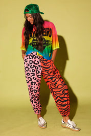 Model wearing a colorful oversized mesh tee, pink and orange patterned jogger pants with hands in pockets, green trucker hat, and sneakers against a pale yellow background