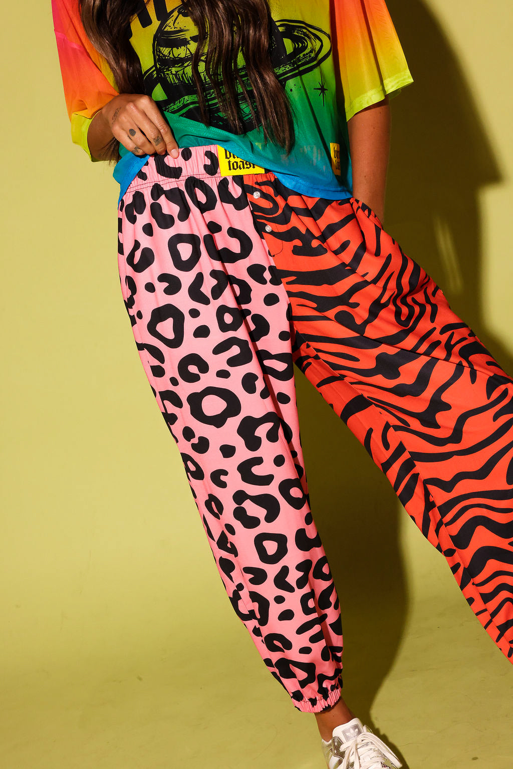 Model holding the waistband, wearing a colorful oversized mesh tee, pink and orange patterned jogger pants, and sneakers against a pale yellow background.