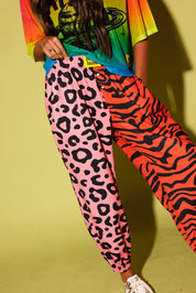 Model holding the waistband, wearing a colorful oversized mesh tee, pink and orange patterned jogger pants, and sneakers against a pale yellow background.