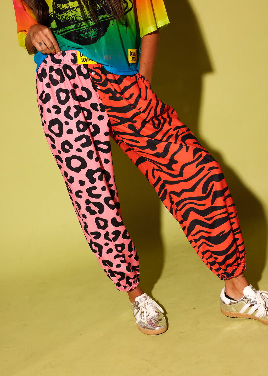 Close up of a model wearing a colorful oversized mesh tee, pink and orange patterned jogger pants, and sneakers against a pale yellow background