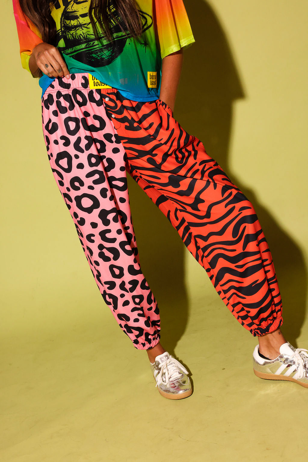 Close up of a model wearing a colorful oversized mesh tee, pink and orange patterned jogger pants, and sneakers against a pale yellow background