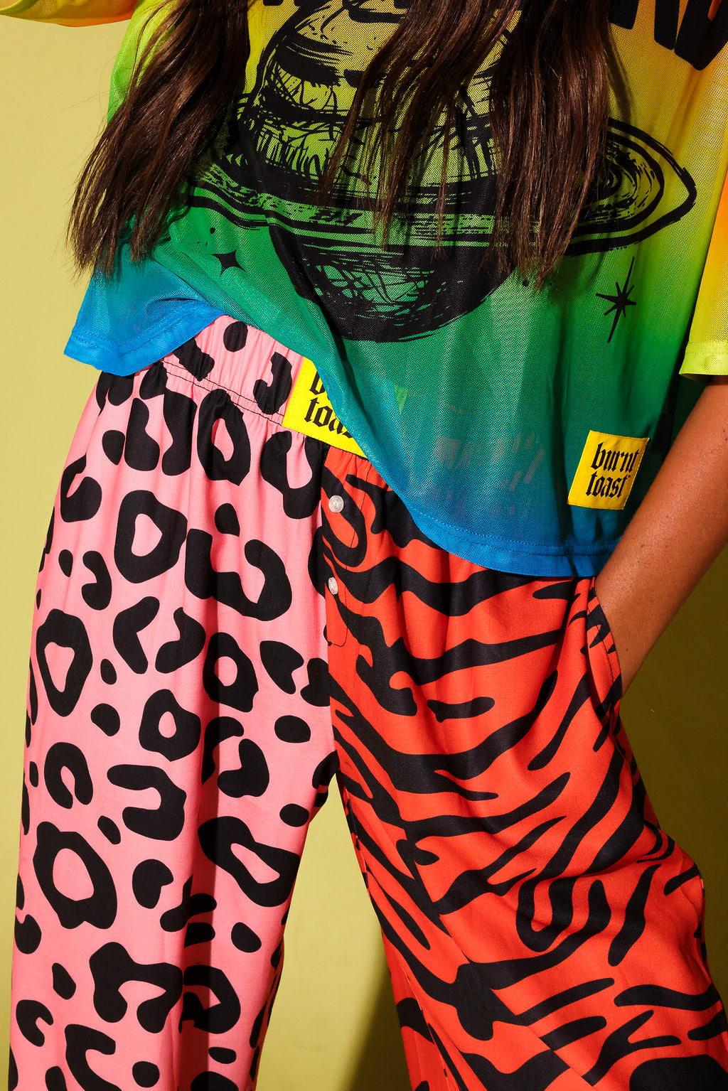 Close up of a waistband of pink and orange patterned jogger pants and yellow burnt toast tag, a colorful oversized mesh tee, against a pale yellow background.