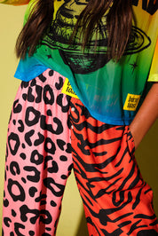 Close up of a waistband of pink and orange patterned jogger pants and yellow burnt toast tag, a colorful oversized mesh tee, against a pale yellow background.