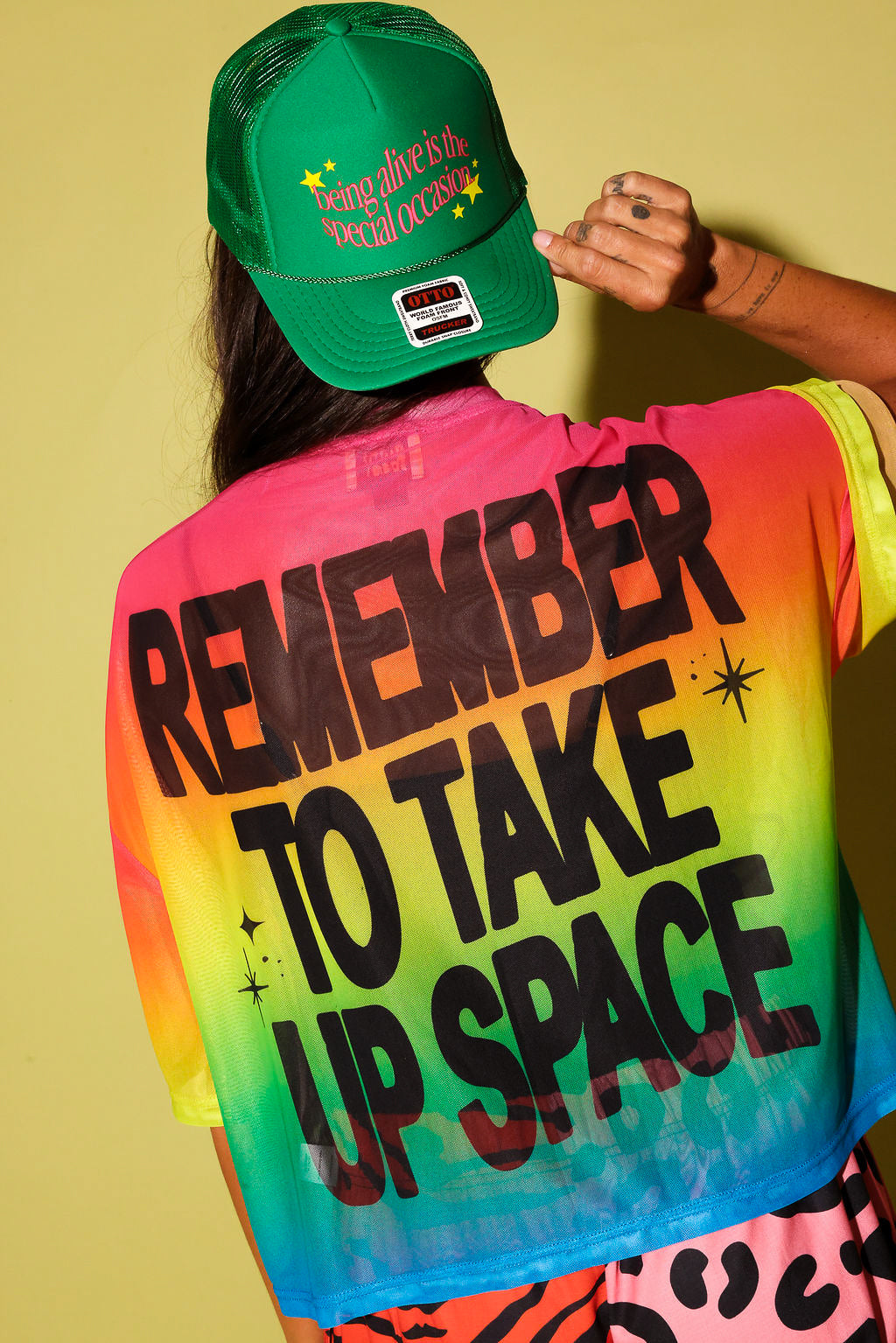 The back of a model wearing a rainbow gradient mesh top with black text that reads 'REMEMBER TO TAKE UP SPACE' and a green trucker at, on a pale yellow background