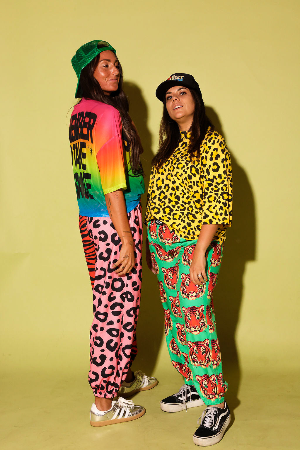 Two models wearing colorful outfits, the pants are loose fitting and the tops are cropped short sleeve, and trucker hats against a pale yellow background