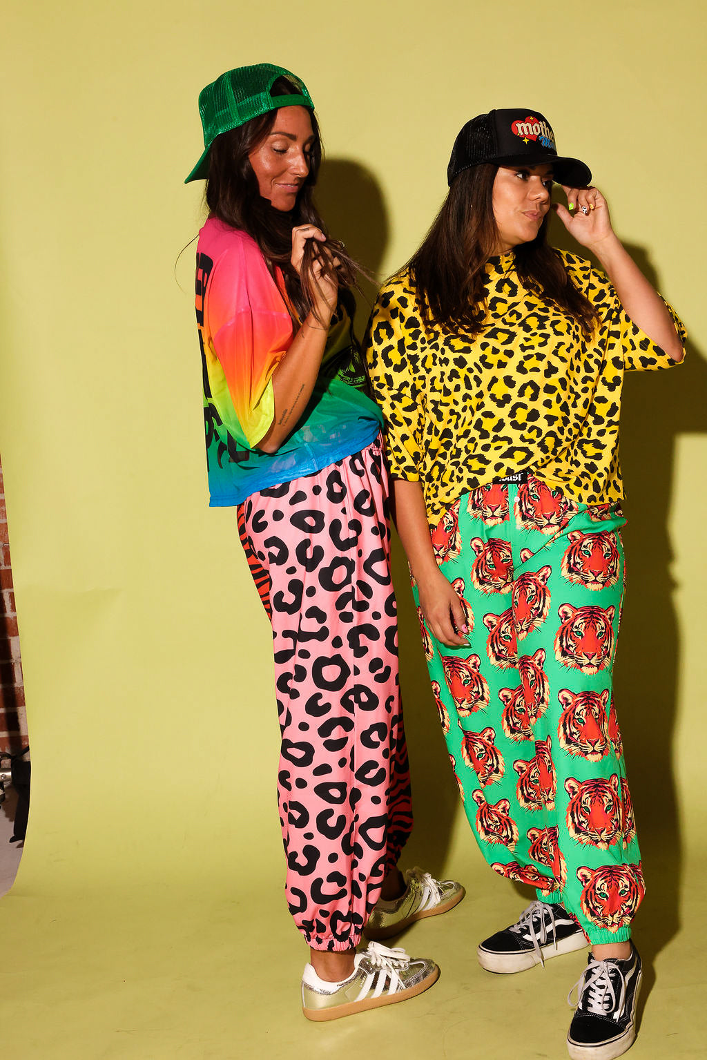 Side view of two models wearing colorful outfits that include mesh tops, hats, and sneakers, on a pale yellow background