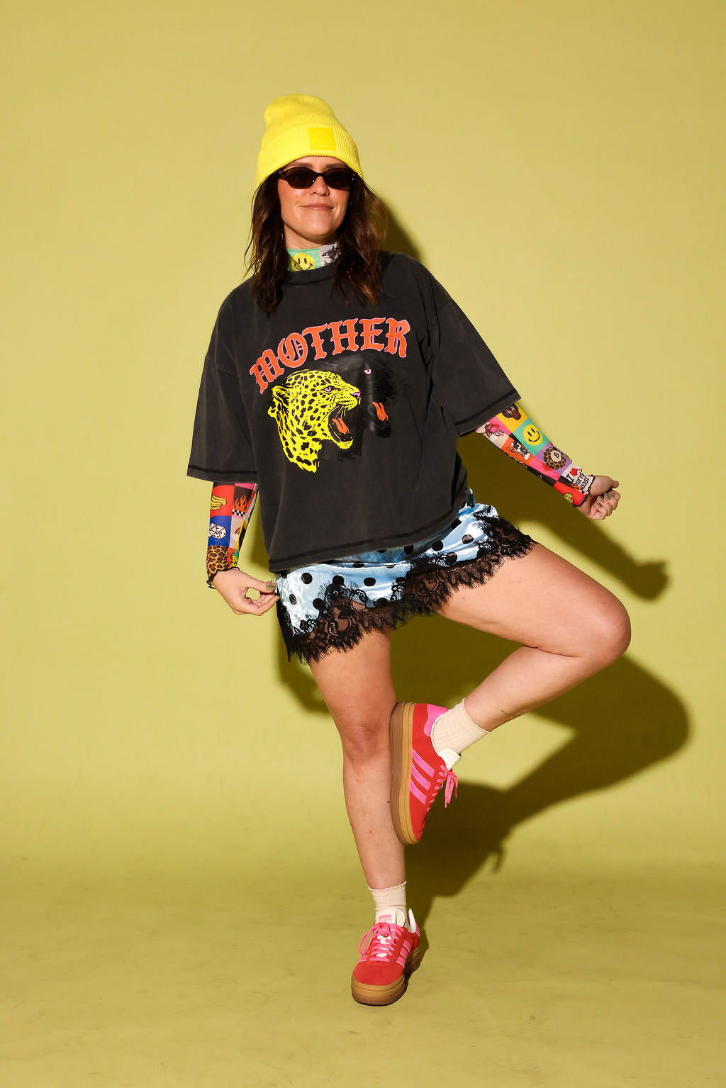 Model wearing a grey graphic tee with the word 'MOTHER' in pink and two panthers and a blue and black polka dot lace slip dress and pink and yellow adidas on a pale yellow background