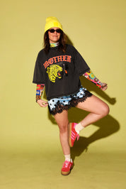 Model wearing a grey graphic tee with the word 'MOTHER' in pink and two panthers and a blue and black polka dot lace slip dress and pink and yellow adidas on a pale yellow background