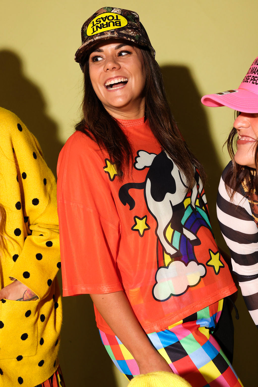 A woman wearing a bright orange graphic T-shirt with a cow and rainbow design, a camouflage cap with a yellow logo, and colorful striped pants, standing against a yellow wall. Two other people are partially visible on both sides.