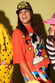 A woman wearing a bright orange graphic T-shirt with a cow and rainbow design, a camouflage cap with a yellow logo, and colorful striped pants, standing against a yellow wall. Two other people are partially visible on both sides.