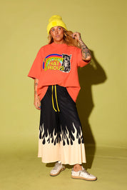 A model stands against a solid olive green studio background wearing a bright orange graphic t-shirt with a rainbow and tiger print, black wide-leg pants with a cream flame-like hem and yellow drawstrings, a yellow beanie, and white sneakers.