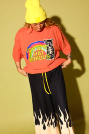 A person wearing a bright yellow beanie, a coral-orange graphic T-shirt with a rainbow and cat print reading 'IT'S NOT THAT SERIOUS', and black wide-leg pants with a yellow drawstring and flame-like cream sections near the hem, posed against a yellow-green studio background.