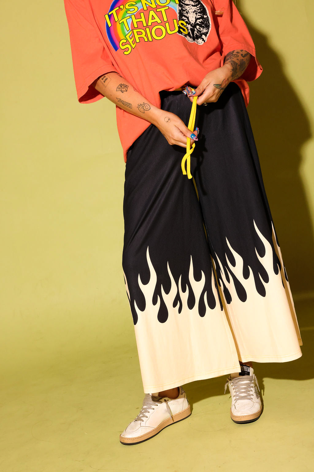 A person wearing wide-leg black pants with a cream flame-like gradient at the hem, styled with an orange graphic t-shirt and white sneakers against a yellow-green studio background.