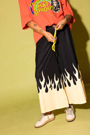 A person wearing wide-leg black pants with a cream flame-like gradient at the hem, styled with an orange graphic t-shirt and white sneakers against a yellow-green studio background.