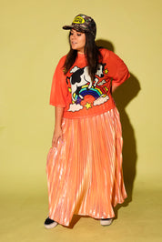 A model wearing an orange mesh top with a cow jumping over a rainbow graphic, a peach metallic plisse skirt and camo burnt toast trucker hat, on a pale green background