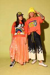 Two women posing together in matching orange graphic t-shirts and wide-leg pants against a solid pale yellow-green studio backdrop. The woman on the left wears a shiny peach pleated skirt, a black cap, and black-and-white sneakers.