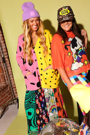 Two women posing in vibrant, colorful outfits. The woman on the left wears a pink cardigan with black spots, a purple beanie, and a multicolor patterned skirt.