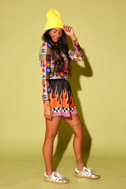 A model wearing a colorful graphic long-sleeve crop top, a black and orange flame-print mini skirt with a pink ruffle hem, a bright yellow beanie, and white sneakers, standing against a solid pale yellow-green studio background.
