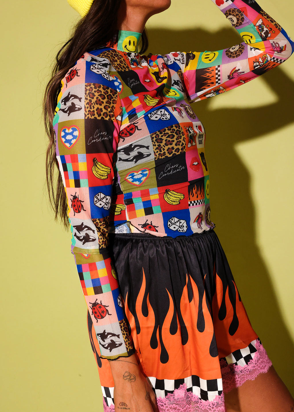 A model wearing a vibrant, multicolored patchwork long-sleeve top with cartoon and animal prints, paired with a black skirt featuring orange flames and pink lace trim, set against a solid yellow-green background.