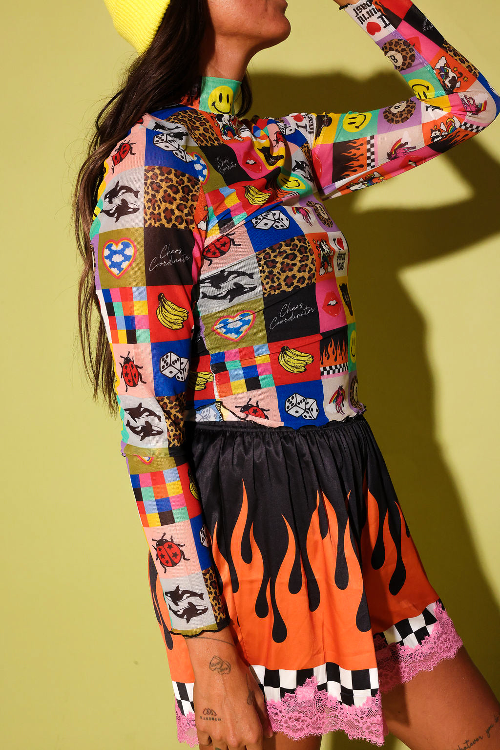 A model wearing a vibrant, multicolored patchwork long-sleeve top with cartoon and animal prints, paired with a black skirt featuring orange flames and pink lace trim, set against a solid yellow-green background.
